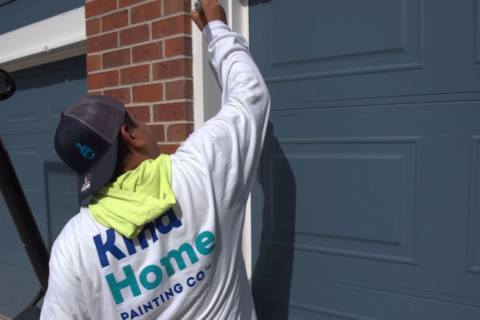 A Kind Home painter in branded gear cutting in white trim around a garage door